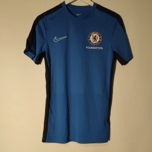 Chelsea fotbollströja från Nike - Säljer en blå Chelsea fotbollströja från Nike med svarta detaljer längs axlar och sidor. Tröjan har korta ärmar och rund hals. Perfekt för träning eller match och har Chelsea-logga på bröstet. Fick den under ett läger i England därav foundation under loggan. Storlek S. Kolla gärna in min profil för andra fotbollströjor och grisch kläder.