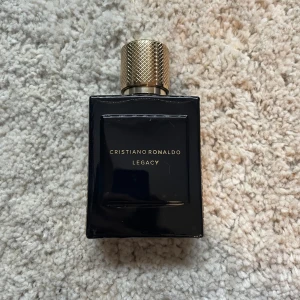 Cristiano Ronaldo Legacy Eau de Toilette - Säljer en elegant herrparfym, Cristiano Ronaldo Legacy. Flaskan är svart med guldiga detaljer och har en lyxig, fyrkantig design. Lockets ovansida pryds av CR-loggan. Perfekt för dig som gillar exklusiva dofter. ungefär 75ml kvar och har inte original paketet men den är äkta