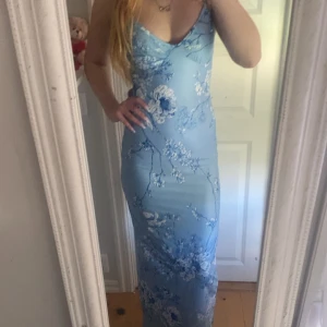 Ljusblå blommig långklänning - Säljer en ljusblå långklänning med v-ringning och blommigt mönster. Klänningen har en tight passform och är ärmlös, perfekt för sommarens tillfällen. Materialet känns mjukt och följsamt mot kroppen. 
