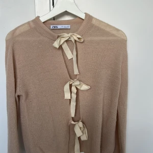 Beige kofta med rosetter från Zara - Säljer en beige kofta från Zara med långa ärmar och tre dekorativa rosetter framtill istället för knappar. Koftan är tunnstickad och har en mjuk känsla, perfekt för lager-på-lager. Enkel och stilren design som passar till mycket. Använd enstaka gånger, så som ny!! (Kan anses som lite rosa i nyansen)