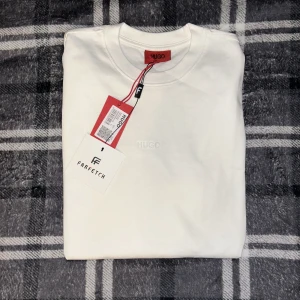 Vit t-shirt från Hugo - Säljer en stilren vit t-shirt från Hugo med diskret logga framtill. Klassisk rund halsringning och mjukt bomullsmaterial. Perfekt basplagg för garderoben. T-shirten är märkt med både Hugo och Farfetch-taggar.