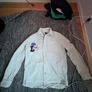 Vit skjorta med tryck och patchar - Vit skjorta från Q/S designed by med tryck på ryggen i blått och rött samt texten 'Legendary mood'. Framsidan har patchar och bröstficka. Klassisk krage och knäppning framtill.