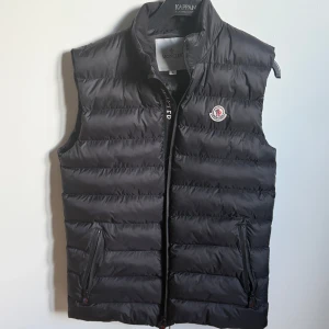 Svart dunväst från Moncler - Snygg svart dunväst från Moncler med klassisk logotyp på bröstet. Västen har hög krage, dragkedja framtill och två fickor med dragkedja. Perfekt för lager-på-lager och stilren look.