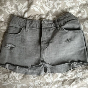 Grå jeanskjol med slitningar - Säljer en grå jeanskjol med rå kant och slitna detaljer framtill. Kjolen har klassiska fem fickor och bälteshällor. Kort modell som passar perfekt till sneakers eller boots.