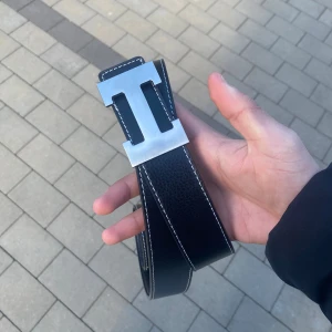 Svart bälte med silverfärgat H-spänne - Säljer ett svart bälte med tydliga vita sömmar och ett stort, stilrent H-format spänne i silverfärg. Bältet har en klassisk design och är gjort i skinn. Perfekt accessoar för att lyfta din outfit.