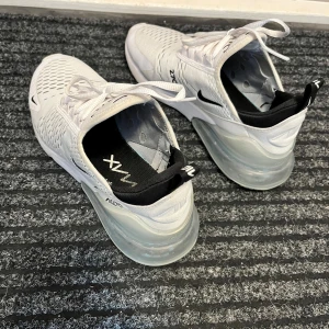 Vita Nike Air Max 270 sneakers - Säljer ett par fräscha vita Nike Air Max 270 sneakers med svart detalj och stor synlig luftbubbla i hälen. Skorna har snörning, ventilerande mesh och en sportig look. Väldigt bra skick!