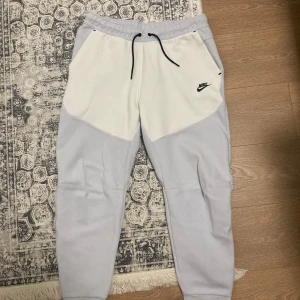 Nike tech fleece  - Nike mjukisbyxor i vitt och ljusgrått med svarta snören och logga på vänster sida. Byxorna har elastisk midja, snörning och fickor fram samt en bakficka med dragkedja. Perfekta för en avslappnad stil.