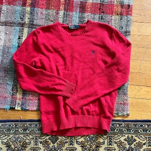 Röd stickad tröja från Polo Ralph Lauren - Säljer en klassisk röd stickad tröja från Polo Ralph Lauren med rund halsringning och blå broderad logga på bröstet. Tröjan har långa ärmar och ribbade muddar vid ärmslut och nederkant. Perfekt för lager-på-lager-stil.