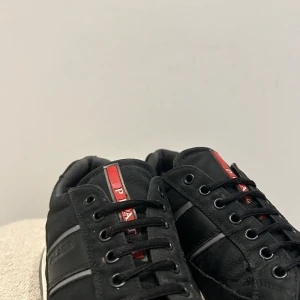 Svarta sneakers från Prada med röda detaljer - Säljer ett par svarta sneakers från Prada med röda och grå detaljer. Klassisk Prada-logga syns på plösen och innersulan. Perfekta för dig som gillar stilrena och exklusiva sneakers.