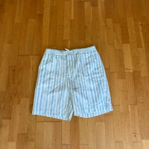 Randiga blå och vita shorts från J.Lindeberg - Säljer ett par fräscha blå- och vitrandiga shorts från J.Lindeberg. De har elastisk midja med snörning och är tillverkade i bomull. Perfekta för sommaren med sin lätta och luftiga känsla.