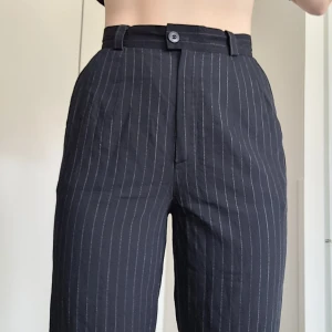 Svarta kostymbyxor med vita ränder - Säljer ett par svarta kostymbyxor med tunna vita ränder. Byxorna har hög midja, raka ben och stängs med knapp och dragkedja. Klassisk passform med bälteshällor och diskret randigt mönster. Perfekta för en stilren look.