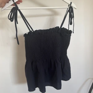 Svart smockad linnetopp med volang - Supersöt svart linnetopp med smockad överdel och volangkant upptill. Axelbanden är tunna och knyts upptill för en justerbar passform. Perfekt till varma dagar och enkel att matcha med jeans eller kjol.