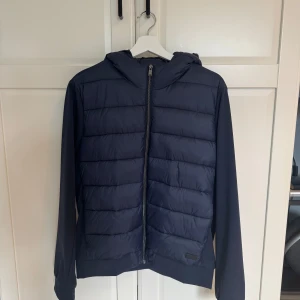 Mörkblå pufferjacka med huva från SOCI3TY - Snygg mörkblå pufferjacka från SOCI3TY med huva och dragkedja framtill. Jackan har quiltad framsida och släta ärmar, perfekt för kyliga dagar. Enkel och stilren design som passar till mycket. Använd 3-4 gånger. PM vid frågor 