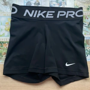 Svarta Nike Pro shorts - Svarta Nike Pro shorts med bred resår i midjan och stor logga framtill. Tight passform och korta ben, perfekta för träning eller sport. Tillverkade i syntetmaterial som andas och ger bra rörelsefrihet. Använd få tal ggr, 2-3 ggr. Liten repa på baksidan ( sista bilden) men syns inte alls! (Är alltså inget hål utan bara en repa!) Säljer pga de var för små. Hör av dig vid frågor! 🤩