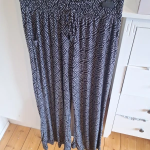Svartvita vida byxor med grafiskt mönster - Säljer ett par luftiga svartvita byxor med grafiskt diamantmönster. Byxorna har resår i midjan och är tillverkade i mjuk polyesterblandning med lite stretch. Perfekta för en avslappnad och trendig look.