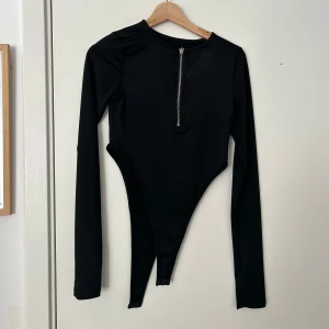 Svart långärmad bodysuit med dragkedja - Svart långärmad bodysuit med rund hals och en snygg dragkedja framtill. Tight passform och högt skurna ben. Perfekt att styla med jeans eller kjol för en trendig look.