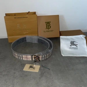 Rutig Burberry bälte med silverdetaljer (Aldrig Använt) - Säljer ett klassiskt bälte från Burberry med det ikoniska rutiga mönstret i beige, svart och rött. Bältet har ett stort silverspänne med Burberry-logga och är tillverkat i skinn. Perfekt accessoar för att lyfta din outfit.