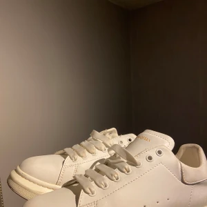 Vita sneakers från Alexander McQueen - Säljer ett par klassiska vita sneakers från Alexander McQueen med tjock sula och diskret guldfärgad logga på hälen. Skorna har rund tå och snörning, perfekta för en stilren look.