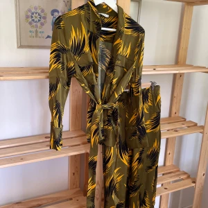 Grön kostym med grafiskt mönster från Samsoe - Säljer en tvådelad kostym med kavaj och vida byxor i grönt med gult och svart grafiskt mönster. Kavajen har bälte i midjan som man ta av. Lite sönder i trådarna på höger ben. S på överdel, M på byxorna