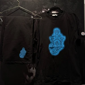 Svart t-shirt och shorts från Moncler med blått tryck - Svart t-shirt och shorts från Moncler med ett stort blått tryck på bröstet och Moncler-logga på vänster ärm. Klassisk rund halsringning och normal passform. Perfekt för dig som gillar streetwear och vill sticka ut med en snygg detalj.
