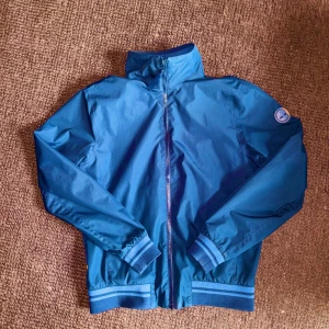 Timberland jacka i perfekt skick - Snygg blå jacka från Timberland med dragkedja framtill och ribbade muddar vid ärmslut och nederkant. Jackan har Timberland-logga på ärmen. Vindtät och vattenavvisande. Mesh-foder och bra innerficka. Köpt i Timberlandbutiken 2023 för 1799kr. Sparsamt använd och i perfekt skick.