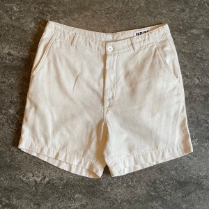 Vita Van Shorts från HOPE - Stilrena vita shorts från HOPE i modellen Van Shorts. Klassisk design med snedställda fickor fram, en bakficka med knapp och gylf med dragkedja. Perfekta för varma dagar och enkel att matcha med olika outfits.