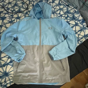 Blå och grå vindjacka från Under Armour - Säljer en snygg tvåfärgad vindjacka från Under Armour i ljusblått och grått. Jackan har huva, dragkedja framtill och två fickor med dragkedja. Perfekt för blåsiga dagar och enkel att matcha med det mesta.