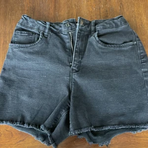 Svarta jeansshorts från Name It - Svarta jeansshorts med fransig kant från Name It. Klassisk femficksmodell med bälteshällor och dragkedja. Hög midja och normal passform, perfekta för sommaren.
