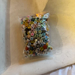 Färgglada bokstavspärlor för smyckestillverkning - En påse med runda pärlor i olika färger med bokstäver på. Perfekta för att skapa personliga armband, halsband eller andra accessoarer. Pärlorna har vit, gul, blå, rosa, svart och grön bakgrund med svarta eller färgade bokstäver.