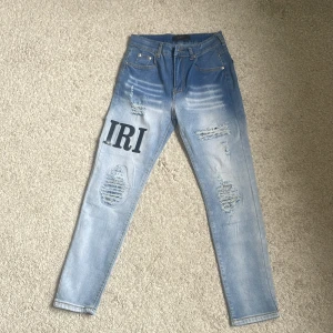 Ljusblå slitna jeans från Amiri - Säljer ett par ljusblå jeans från Amiri med slitningar och tryckta bokstäver på benen. Jeansen har fem fickor, dragkedja och smal passform. Perfekta för dig som gillar streetstyle och unika detaljer.