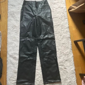 Svarta raka byxor i skinnimitation - Snygga svarta byxor i skinnimitation med raka ben och vita kontrastsömmar. Klassisk femficksmodell med dragkedja och knapp framtill. Perfekta för dig som vill ha en edgy vibe till din outfit.