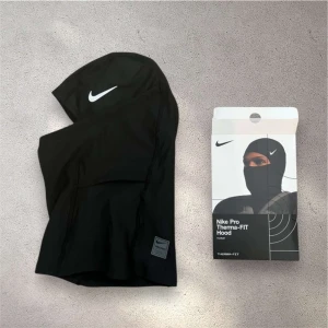Svart Nike Pro Therma-FIT balaclava - Svart balaclava från Nike Pro med Therma-FIT-teknologi. Huvan har en vit Nike-logga framtill och en liten Nike Pro-etikett nedtill. Perfekt för kalla dagar och sportaktiviteter utomhus.