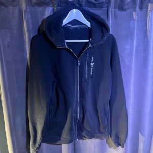 Svart hoodie med dragkedja från sail racing - Säljer en svart hoodie från Peak Performance med dragkedja framtill och huva. Tröjan har en diskret logga på bröstet och en liten logga på huvan. Perfekt för en avslappnad stil.