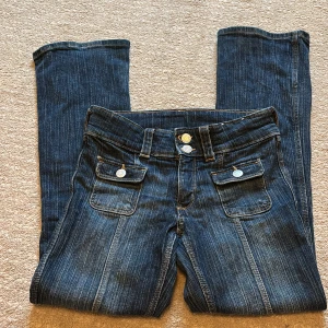 Mörkblå bootcut jeans med fickdetaljer - Säljer ett par mörkblå jeans med bootcut passform och dekorativa fickor framtill med knappar. Jeansen har låg midja och klassiska fickor. Använda få gånger+säljer för de är för små🥰 de finns även inte o köpa i butik längre.