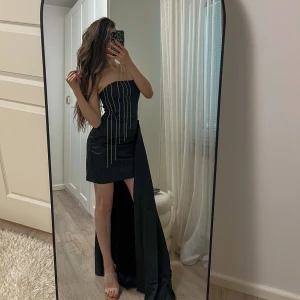 Svart strapless klänning med släp - Supersnygg svart strapless klänning med kort framsida och långt släp på sidan. Dekorerad med glittriga kedjor som hänger från bysten och ger en lyxig vibe. Sitter tight och har en elegant, asymmetrisk look. Perfekt för dig som vill sticka ut på festen!💕Storlek: XS/S