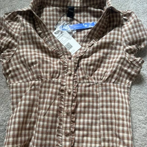 Brun och vit rutig skjorta från Cider - Supersöt brun och vit rutig skjorta från Cider med korta puffärmar, v-ringning och knappar framtill. Skjortan har volangdetaljer längs knappraden och är helt oanvänd (lappen är kvar).