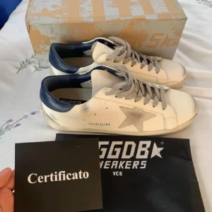 Golden Goose vita sneakers med blå detalj - Golden Goose sneakers i vitt skinn med grå snörning, ikonisk stjärndetalj på sidan och mörkblå häl. Platt sula och rund tå. Snyggt slitna detaljer för en cool look. Kommer med originalpåse och certifikat.