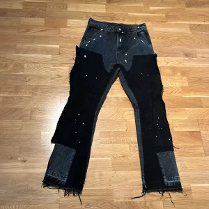 NVLTY Flared jeans - Unika svarta och mörkgrå jeans med patchwork-design, råa och fransiga benslut samt vita färgstänk. Byxorna har flera fickor, dragkedjedetaljer och en loose fit. Perfekt för dig som gillar streetstyle och vill sticka ut.