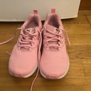Rosa sneakers från Puma strl 38 - Snygga ljusrosa sneakers från Puma med vit sula och svart logga. Skorna har snörning, meshdetaljer och mjuk SoftFoam+ innersula för extra komfort. Perfekta för dig som vill ha en sportig och trendig look.