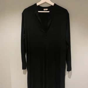 Svart långärmad klänning från Filippa K - En stilren svart klänning från Filippa K med lång ärm och v-ringad hals. Klänningen har en rak passform och är tillverkad i ett mjukt, lätt material som faller snyggt. Perfekt för dig som gillar minimalistisk och tidlös stil. Knälång och med slitsar på sidorna. 