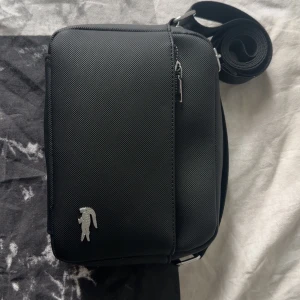 Lacoste axelväska  - Snygg svart axelväska från Lacoste med justerbar rem och silverfärgad dragkedja. Väskan har en liten logga i silver på framsidan och ett extra fack med dragkedja på baksidan och framsidan samt två inre mindre fack. Perfekt storlek för det viktigaste och enkel att bära med sig.