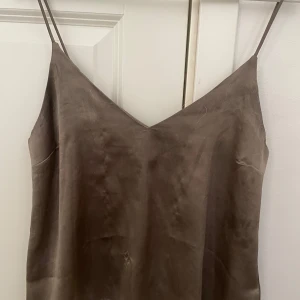 Brun satintopp - Snygg brun satintopp med tunna axelband och v-ringning. Klänningen har en mjuk satinliknande yta som ger en lyxig känsla och faller fint längs kroppen. Perfekt för dig som gillar minimalistisk och stilren look.