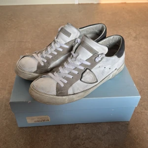 Vita och blå sneakers från Philippe Model - Säljer ett par snygga sneakers från Philippe Model i vitt och blått med blåa detaljer på hälen. Skorna har snörning, rund tå och platt sula. Perfekta för dig som gillar stilrena och klassiska sneakers med en stilig touch.