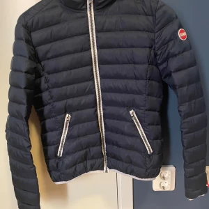Mörkblå pufferjacka från Colmar - Säljer en mörkblå pufferjacka från Colmar med silvriga dragkedjor framtill och på fickorna. Jackan har en kortare passform och quiltad design. Perfekt för kyligare dagar och enkel att matcha med olika outfits.