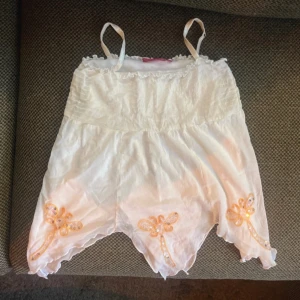 Vit linnetopp med paljett-detaljer  - Supersöt vit linnetopp från KappAhl med smala axelband och volangkant upptill. Nedtill finns broderade trollsländor i orange med paljetter som glittrar. Toppen har en luftig känsla och är perfekt för varma dagar. Passar även XS