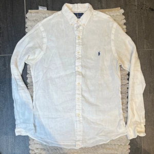 Ralph Lauren Linneskjorta Vit - Stilren linneskjorta i vitt från Ralph Lauren. Klassisk modell i 100 % linne – perfekt för sommar och varmare dagar. Mycket fint skick, endast lite gulnad under armarna, inget som stör användningen. 🔹 Storlek: M 🔹 Material: 100 % linne 🔹 Skick: Mycket fint, mindre missfärgning under armarna  Sökord:  Liknande märken: 
