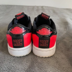 Nike Air Jordans Low Slip-On i svart och rött - Säljer ett par Nike Air Jordans Low Slip-On i svart och rött med vit sula. Skorna har en klassisk Jordan-logga bak och på plösen, samt en stilren design utan snörning. Perfekta för dig som vill ha en ikonisk sneaker med enkel påtagning.