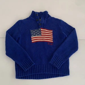 Blå stickad tröja med amerikansk flagga från Polo Ralph Lauren - Säljer en blå stickad tröja från Polo Ralph Lauren med amerikansk flagga framtill och RL67-broderi. Tröjan har hög krage med knappar och långa ärmar. Perfekt för dig som gillar klassisk och sportig stil. Pris kan diskuteras 