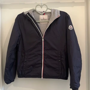 Mörkblå Moncler windbreaker stl. 152cm -  mörkblå vindjacka från Moncler med grå huva och innerfoder. Jackan har dragkedja framtill med röd, vit och blå detalj samt Moncler-logga på ärmen. Den är såklart äkta och i mycket bra skick (nästan oanvänd). Den är i storlek 152cm och 12 år. Vid snabb affär kan pris diskuteras!