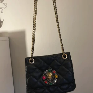 Svart quiltad axelväska från ALDO - Snygg svart quiltad axelväska från ALDO med guldfärgad kedja och dekorativt lejonhuvud med färgglada stenar på framsidan. Väskan har en klassisk form och passar perfekt för att lyfta din outfit.